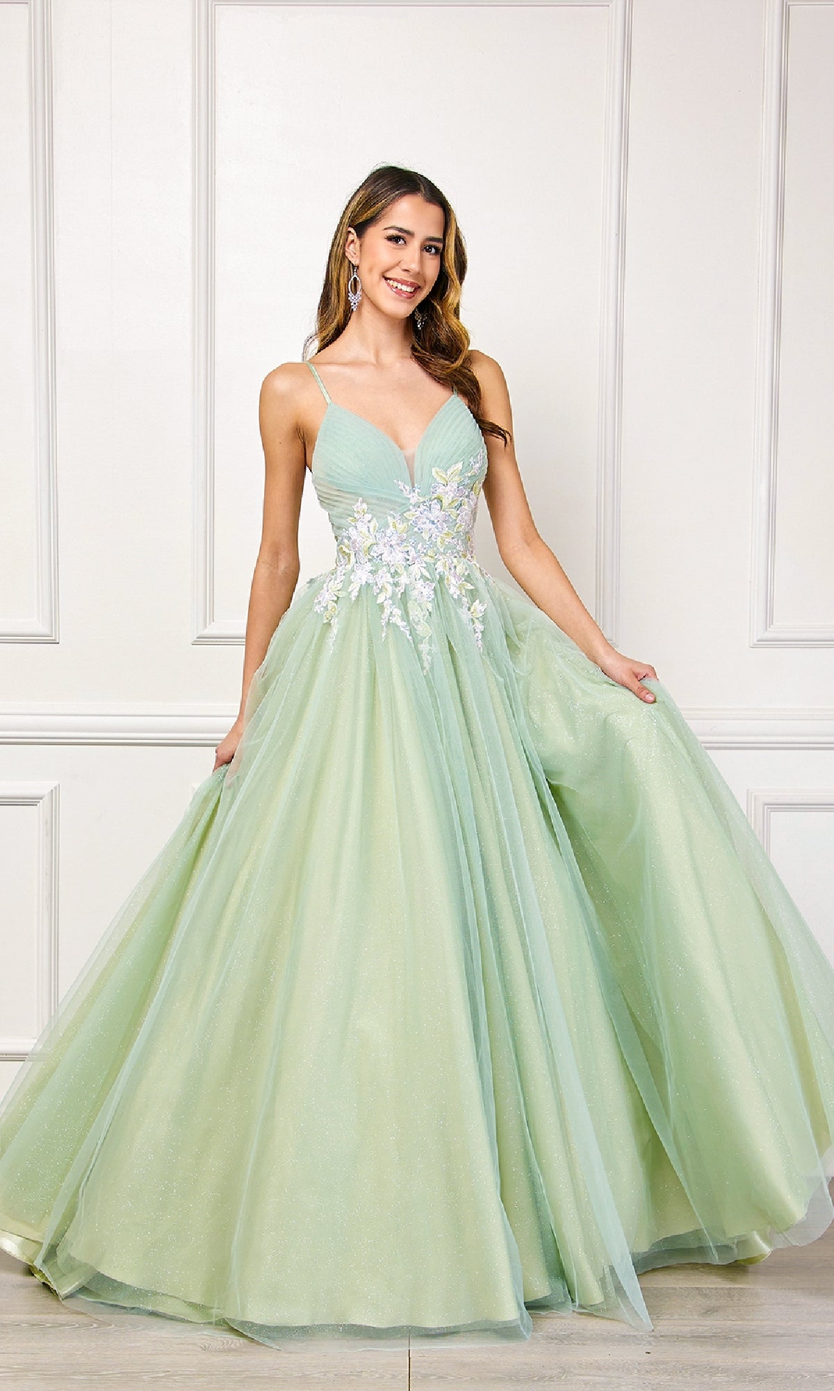 A woman wearing the Long Pastel Prom Ball Gown: Amelia Couture SU088, a light green floor-length A-line dress with floral embellishments, poses against a white paneled wall, holding out her skirt.